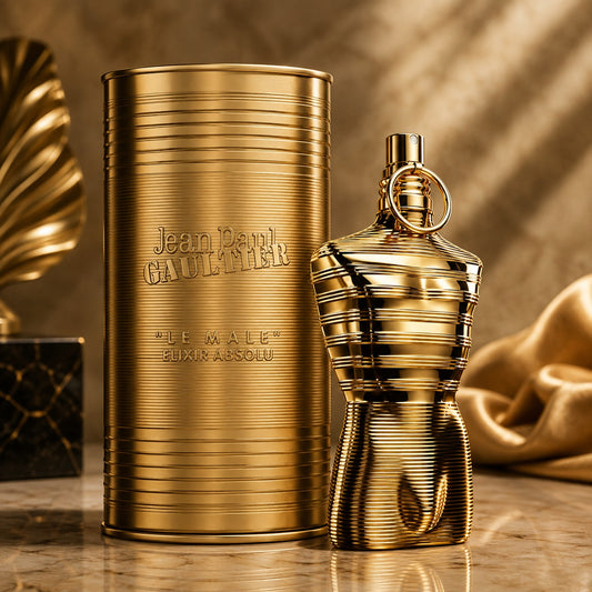Jean Paul Gaultier Le Male Lux Elixir Absolu perfume bottle and matching cylindrical packaging, both in a polished gold finish, displayed on a marble surface with draped fabric in the background.